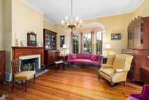 A traditional living room with wooden floors, antique furniture, a fireplace, chandelier, and a large bay window with views outside. The room has pale yellow walls and various wooden cabinets and chairs.