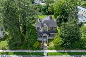 Aerial view of a two-story house with a covered front porch and gabled roof, surrounded by mature trees and greenery, located in a residential neighborhood. Concrete walkway leads to the front steps.