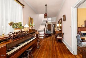 A hallway with polished wooden floors, an ornate upright piano, decorative lamps, and a staircase at the end. Antique furniture and a mirror line the walls, and an open doorway leads to another room.