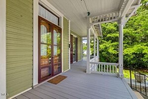 A covered front porch with light gray flooring, green wooden siding, white railings, and trim. Double wooden doors with glass panels open onto the porch, which overlooks green trees and a yard.