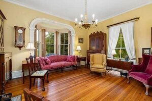 A traditional living room with yellow walls, wood floors, antique furniture including a red settee, yellow armchair, red wingback chair, grandfather clock, wooden cabinet, lamps, large windows, and a chandelier.