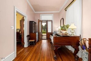A hallway with polished wooden floors, a large wooden piano, chairs, a mirror, lamps, and a doorway leading outside. Light pink walls and tall curtains frame the space, with natural light coming through the glass door.