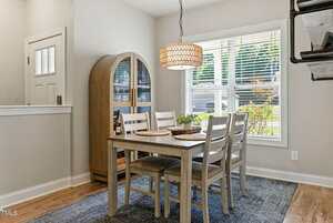 A dining area with a wooden table set for four, matching chairs, a woven pendant light, a wooden cabinet, a window with blinds, and a blue rug on a hardwood floor.