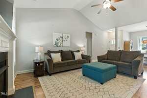 A living room with two dark sofas, a turquoise ottoman, a patterned rug, a side table with a lamp, and a ceiling fan. A piece of wall art hangs above the sofa, and the room opens to a dining area and entryway.