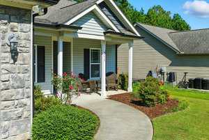 A curved concrete walkway leads to a covered front porch with wicker chairs and a small table. The house has stone and siding exterior, shrubs, and a well-maintained lawn with a neighboring house visible.