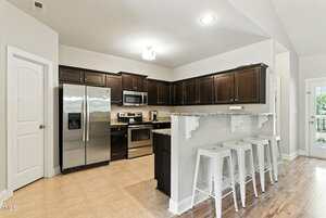 Modern kitchen with stainless steel appliances, dark wood cabinets, granite countertops, and a breakfast bar with three white bar stools. The room has light tile flooring and a white door to the left.