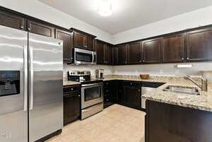 A modern kitchen featuring dark wooden cabinets, stainless steel appliances including a refrigerator, oven, and microwave, granite countertops, a double sink, and beige tile flooring.