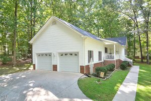 A white single-story house with brick accents, two-car garage, covered front porch, and a concrete driveway, surrounded by trees and green lawn on a sunny day.