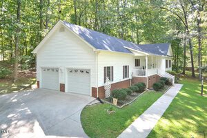 A single-story white house with a dark gray roof, two-car garage, and brick accents is surrounded by trees. A concrete driveway and walkway lead to the covered front porch with white railings.