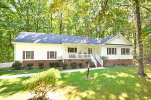 A single-story house with white siding, dark shutters, and a brick foundation. The home has a covered front porch, steps leading up to the entrance, and is surrounded by trees and grass in a wooded setting.