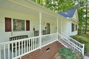 A white house with a covered front porch featuring two black rocking chairs, a porch swing, hanging bird feeders, and white railings. Brick foundation is visible, with green shrubs and trees in the background.
