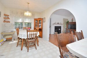 A dining area with a wooden table and chairs, china cabinet, and birdcage. An arched doorway leads to a living room with a fireplace, TV, and more seating. Sunlight comes in through the windows.