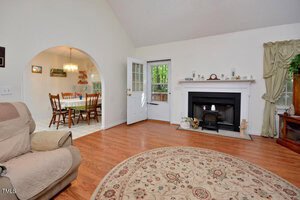 A living room with hardwood floors, a circular patterned rug, a fireplace with a mantel, an open door to the outside, and a view into a dining area with a wooden table and chairs through an arched doorway.