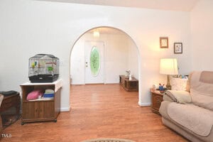A living room with wood floors, a recliner, side tables, a lamp, and a parrot cage on a small table. An arched doorway leads to a foyer with a glass front door and small benches along the walls.