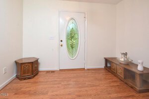 A white door with an oval, decorative green stained-glass window is centered in a room with light-colored walls and wood flooring. Two wooden cabinets and two vases are placed along the walls.