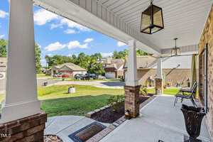 A covered front porch with brick columns overlooks a suburban street with houses, lawns, and parked cars. The porch has two chairs, a potted plant, a doormat, and hanging lantern-style lights.