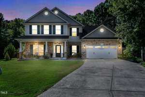 Two-story suburban house with brick and siding exterior, porch, and attached two-car garage. Lights are on inside. Concrete driveway leads to the garage. Trees and grass surround the home at dusk.