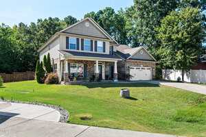 Two-story house with beige siding and brick accents, a covered front porch, and a double garage. The house is surrounded by a well-maintained green lawn and bordered by trees in the background.