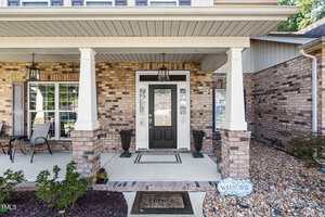 A brick house front porch with two white columns, a dark front door flanked by sidelights, two black lantern lights, a "Home" doormat, potted plants, a welcome sign, and outdoor seating.
