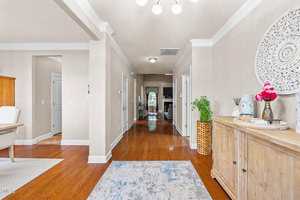 A bright hallway with light beige walls, wooden floors, and white trim. A wooden sideboard with decor sits on the right, and a patterned rug is on the floor. The hallway leads to a living area.