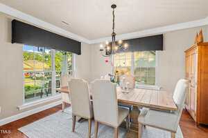 A dining room with a wooden table, six upholstered chairs, a chandelier, large windows with black valances, an orchid plant centerpiece, and an armoire in the corner. Sunlight comes through the windows.