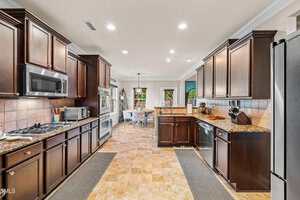 A spacious kitchen with dark wood cabinets, granite countertops, stainless steel appliances, and tan tile flooring, leading into a dining area with a round table and chairs.