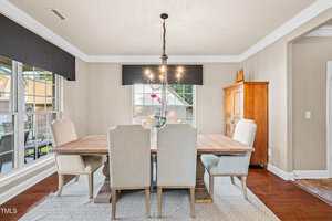 A dining room with a wooden table, six upholstered chairs, a chandelier above the table, large windows on one wall, light-colored walls, a wooden cabinet, and hardwood flooring with a rug under the table.