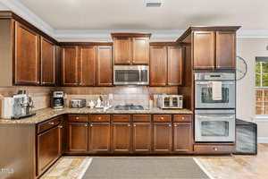 A kitchen with dark wood cabinets, stainless steel appliances including a microwave, stovetop, and double oven, granite countertops, small kitchen appliances, and a window letting in natural light.