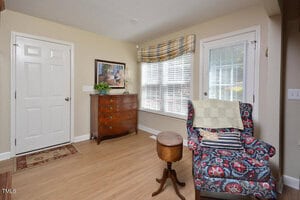 A small sitting area with a patterned armchair, quilt, and round side table next to a window with plaid valance, a wooden chest of drawers, framed art, and a door on light wood flooring.