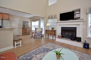 A living room with a fireplace, TV, and two side tables. There is a patterned armchair, wooden bench, and a blue and white rug. Windows have plaid valances, and a kitchen area is partially visible on the left.
