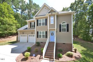 Two-story beige house with black shutters, a blue front door, and a brick foundation. Three-car garage is on the left, with steps and landscaping leading to the entrance. Trees and grass surround the house.