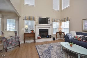 A living room with high ceilings, a fireplace topped with a TV, and large windows. There is a patterned armchair, a couch with pillows and a throw, a coffee table with a plant, and tables with lamps on either side of the fireplace.