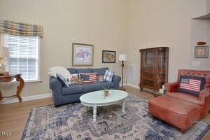 A living room with a blue sofa, a red leather armchair, a patterned rug, a light-colored oval coffee table, wall art, a wooden display cabinet, and a window with a plaid valance. Pillows feature American flag designs.
