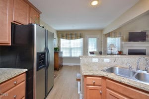 Kitchen with stainless steel refrigerator, granite countertops, and double sink, opening to a living area with large windows, wood flooring, and a fireplace with a TV above the mantle.