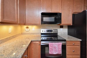A kitchen with light brown cabinets, granite countertops, a black electric stove with a pink towel hanging on the handle, a black microwave above the stove, and a black refrigerator on the right.