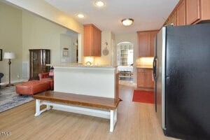 A kitchen with wooden cabinets, stainless steel refrigerator, granite countertop peninsula, and a wooden bench in the foreground. There is a living area with a red lounge chair and rug in the background.
