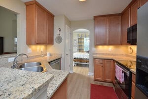 A modern kitchen with granite countertops, wooden cabinets, stainless steel sink, stove, and dishwasher, light wood flooring, under-cabinet lighting, and a red rug, with a view into a bedroom through an open doorway.