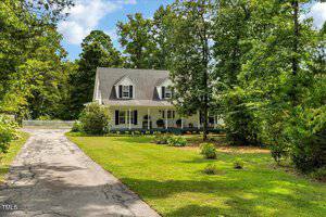 A two-story house with white siding and a covered front porch sits among tall trees and green grass. A paved driveway leads to the house, and a white picket fence is visible in the background.