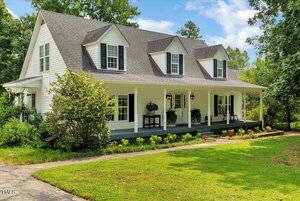 This charming two-story white house at 1711 W B Street, Butner, features a gray roof, three dormer windows, and a spacious covered front porch with black rocking chairs and potted plants, set amid lush green grass, trees, and shrubs.