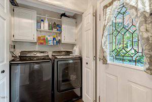 Laundry room with a black washer and dryer, white cabinets above, shelves holding cleaning supplies, and a white exterior door with a decorative glass window and floral curtain. Trees are visible through the door.