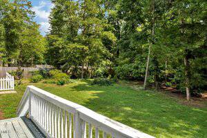 View of a grassy backyard bordered by tall trees and shrubs, seen from a white wooden deck with railing. A wooden fence runs along the back of the yard, and the sky is partly cloudy.