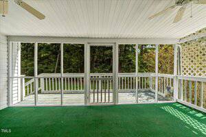 A screened-in porch with a green carpet, white ceiling, and ceiling fan opens to a wooden deck with a railing. Trees and greenery are visible through the screens in the background.
