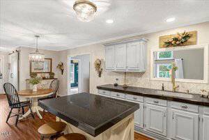 A kitchen with gray cabinets, black countertops, an island with two wooden stools, a window above the sink, and a round dining table with black chairs. Light fixtures hang from the ceiling.