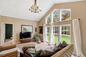 A living room with vaulted ceilings, large windows letting in natural light, a chandelier, neutral walls, a TV on a stand, houseplants, framed art, and cushioned seating arranged around a coffee table.