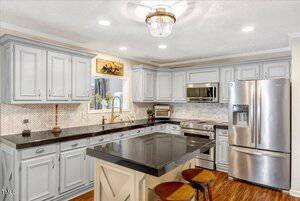A kitchen with light gray cabinets, black countertops, stainless steel appliances, a chevron tile backsplash, a central island with two stools, and wood flooring under recessed and ceiling light fixtures.