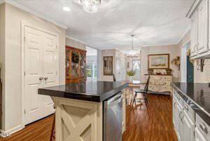 A kitchen with dark countertops, white cabinets, a center island, and wood flooring opens to a dining area with a chandelier, sideboard, chairs, wall art, and decorative shelves. A double-door pantry is on the left.