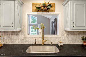A kitchen sink with a gold faucet is set in a black countertop. Light gray cabinets and herringbone tile backsplash surround a window with a decorative wooden flower arrangement above it.