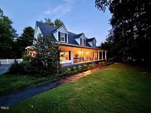 A two-story house with a large wraparound porch, white siding, and dormer windows is illuminated in the evening. The yard has green grass, bushes, trees, and a paved walkway leading to the porch steps.