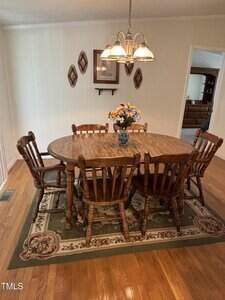 A wooden dining table with six matching chairs sits on a patterned rug. A vase with colorful flowers is centered on the table. Decorative wall art and a light fixture are above. Hardwood floor and neutral walls surround the area.