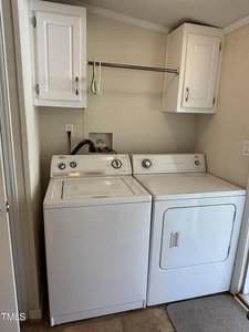 A laundry area with a white washing machine and dryer side by side, white cabinets above, a metal hanging rod, and beige walls. There is a wall outlet and hoses connected behind the machines.